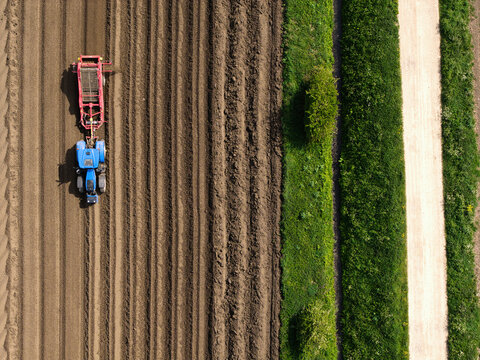 Aerial view of a blue tractor tilling a brown field next to a green hedge and a dirt path in Knottingley, England, United Kingdom.