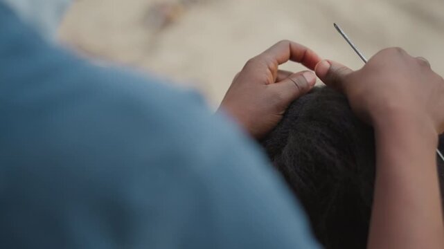 Closeup hands crocheting hair with needle, patient stylist working textured hair into protective rows on outdoor bench, focus on fingers, scalp parting, comb and thread, handcrafted technique, quiet