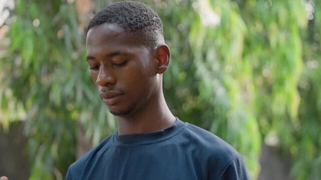 Young black man outdoor portrait contemplative. Downward gaze, soft natural light, beads of sweat on cheek, subtle head tilt across frames showing pause and reflection, casual cotton shirt, blurred