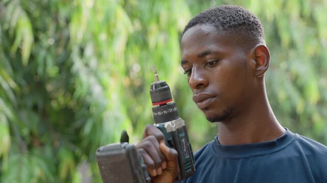 Young man holding drill outdoors, contemplative carpenter testing power tool, beads of sweat on face, leafy backdrop, hands gripping drill, closeup portrait, focused expression, DIY carpentry scene,
