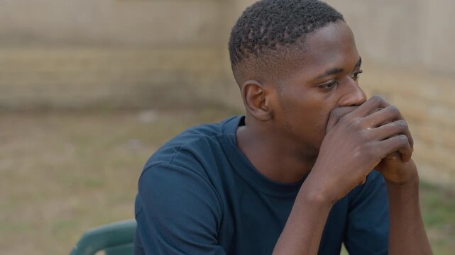 Pensive Black carpenter seated outdoors reflecting on dusty hands and fatigue, quiet yard backdrop, hands clasped near mouth, solemn expression after hard day of carpentry, authentic portrait