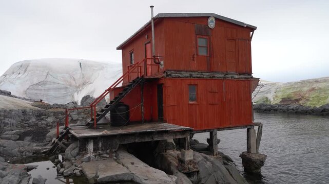Stationary shot of a weathered orange two-story research building at Argentina's Melchior Base, elevated on stilts over rocky Antarctic coastline with a glacier in the background