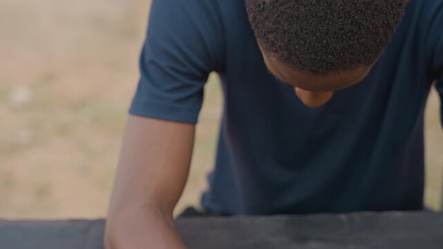 Black apprentice carpenter leaning over bench, overhead view showing torso and forearms coated in sawdust, focused youth inspecting wood surface, quiet outdoor carpentry setting, sunlit texture