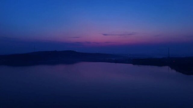 Static aerial view of a calm lake reflecting a deep purple and blue twilight sky with a wind turbine silhouette in China.