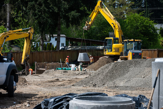 Excavator boom arm being used to lower precast concrete stormwater system access vault into trench, construction workers guiding the installation process
