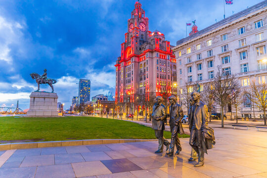 The Beatles Statue stands at Pier Head in front of the illuminated Port of Liverpool Building during twilight.