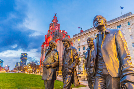 The Beatles Statue stands at Pier Head in front of the illuminated Port of Liverpool Building during twilight.