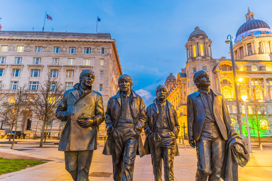 The Beatles Statue stands at Pier Head in front of the illuminated Port of Liverpool Building during twilight.