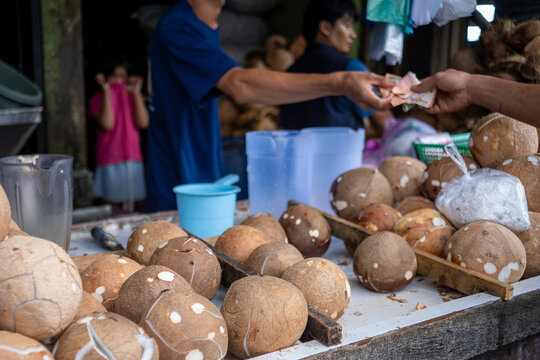 A customer pays with Indonesian rupiah in soft focus at a market stall, with peeled coconuts and grated coconut in the foreground.