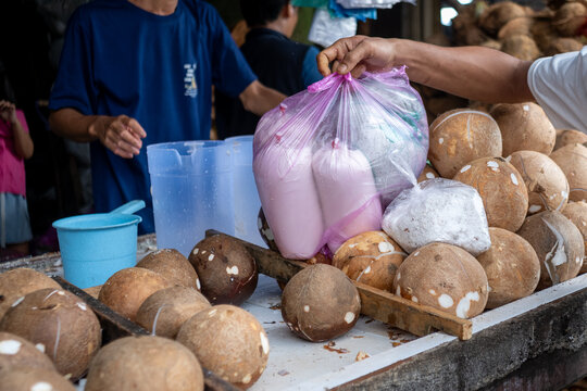 A customer receives bags of fresh coconut cream (santan) at a market stall, with peeled coconuts and grated coconut in the foreground.