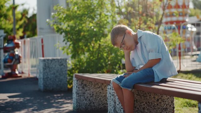 Caucasian boy sitting on bench family park weekend, wearing glasses and denim shorts, head resting on hand, pensive expression, empty amusement rides in background, warm sunlight filtering