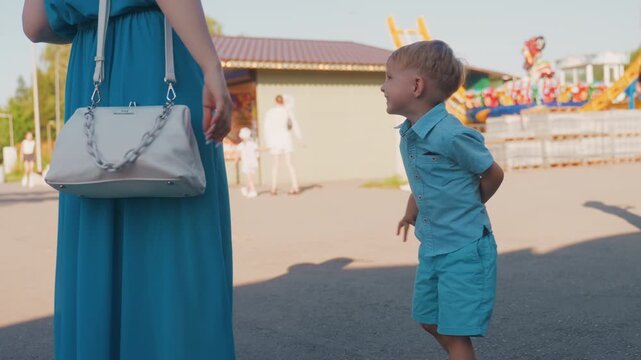 White mother and two boys talking near amusement stalls at sunny park weekend, mother in blue dress holding bag, younger boy pleading while older boy watches, pavement shadows, candid family