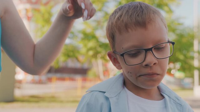Young boy with glasses in park, mother gently pats head while child with freckles shows shy, pensive expressions warm summer sunlight filters through trees, playground blurred in background tender