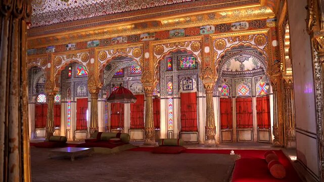 A wide static shot of the Takhat Vilas chamber in Mehrangarh Fort, featuring vibrant stained glass windows, ornate gold-leaf pillars, and traditional floor seating with red cushions, Jodhpur, India