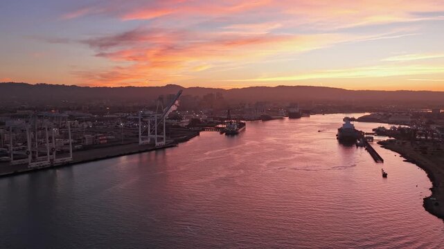 Stunning aerial footage showing the Port of Oakland and the Alameda estuary at dawn, with cargo ships and cranes silhouetted against a beautiful sunrise reflecting on the calm water