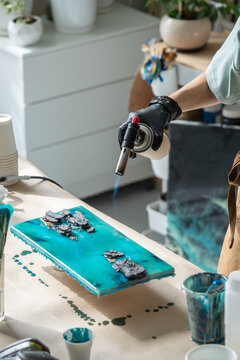 Woman artist in protective gloves using gas torch equipment to create fluid art with epoxy resin, female crafter blowing on canvas with epoxy resin painting, removing air bubbles with torch tool.