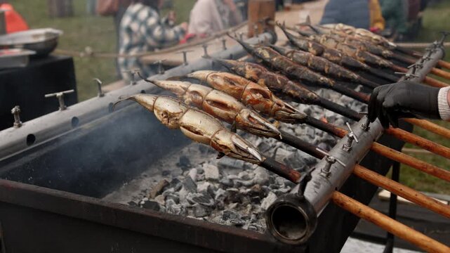 Cook turning fish on a traditional rotisserie grill over charcoal. Street food chef grilling seafood outdoors. Process of preparing roasted fish for meal time.