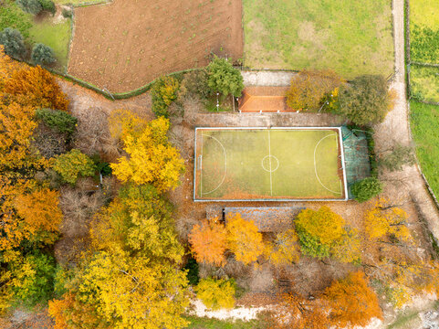 Aerial view of a small soccer pitch surrounded by vibrant autumn trees with yellow and orange foliage in Alvoco das Varzeas, Coimbra, Portugal.