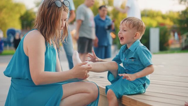 White mother comforting young boy on park boardwalk, applying bandage to scraped knee, barefoot child in denim outfit, summer weekend family atmosphere, gentle bonding under warm light, blurred