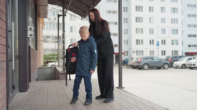 White working mother adjusts sons jacket and inspects backpack at doorstep, zipper and strap check, quick pep talk and gentle guidance, urban parking and apartment facade, preparing for school day