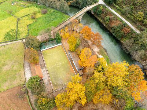 Aerial view of Praia Fluvial de Alvoco das Varzeas featuring a soccer court surrounded by golden autumn trees and a stone bridge over the river in Alvoco das Varzeas, Coimbra, Portugal.