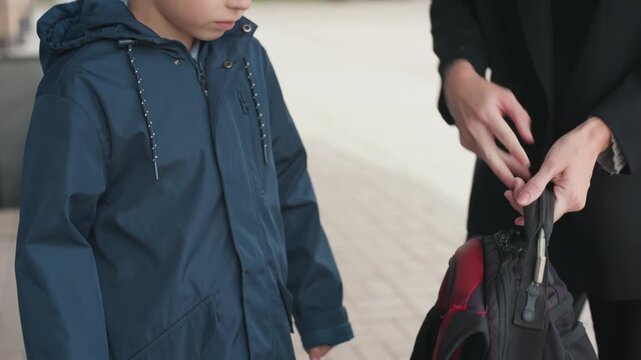 White boy receiving backpack from white woman, hands adjust straps onto shoulders, steadying touch, confident posture, red lined bag detail, morning sidewalk, school readiness, tender goodbye
