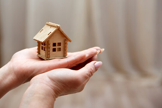 Hands holding a tiny wooden house model. Conceptual image for real estate, mortgage, home insurance, and construction. Soft bokeh background, warm lighting, symbolic gesture of protection and care.
