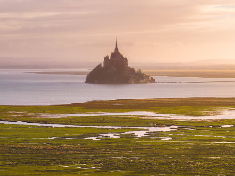 Aerial view of the historic abbey rising above the tidal waters and lush green salt marshes during a soft golden sunset Mont Saint-Michel, Normandy, France.