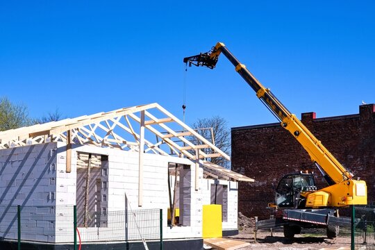 Construction of a modern residential house from aerated concrete blocks. Yellow telescopic crane lifting wooden roof trusses on a building site under clear blue sky.