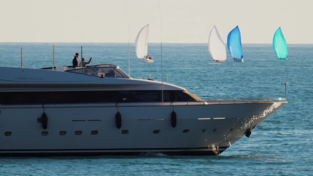 Large white yacht sailing on calm sea with colorful regatta sailboats on horizon during golden hour.