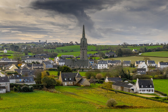 Aerial view of the historic village and its tall gothic church spire surrounded by lush green fields and rolling hills under a dramatic cloudy sky Goulven, Brittany, France.