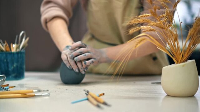 Hands shaping dark clay on workbench with pottery tools near wheat.