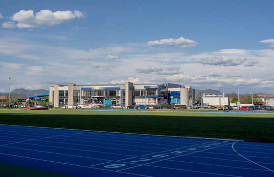 Exterior view of the Aqualand Deva water park complex as seen from the municipal stadium, featuring a professional blue running track in the foreground and the sports base under a bright, cloudy sky.
