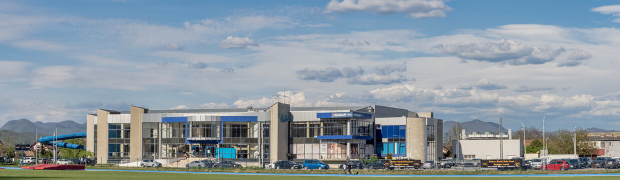 Wide panoramic view of the Aqualand Deva water park complex and the municipal stadium, featuring a professional blue running track in the foreground and a mountain range in the distant background