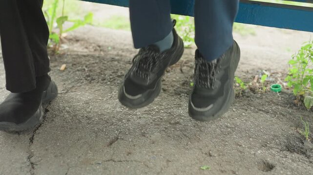 School shoes on cracked pavement waiting, two students sit on blue bench preparing for first day, laces tied, feet tapping, anxious anticipation, subtle greenery at curb, muted morning light