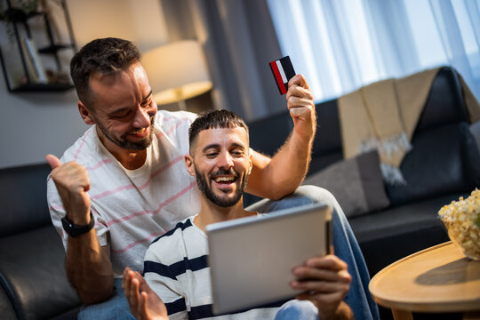 Overjoyed same sex male partners celebrating an approved credit card payment on a laptop at home.