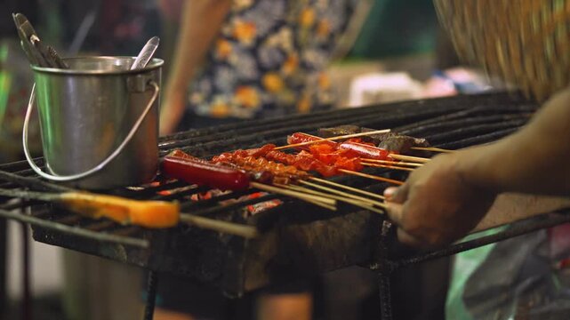 Street BBQ grilling pork skewers while fanning light smoke over charcoal fire