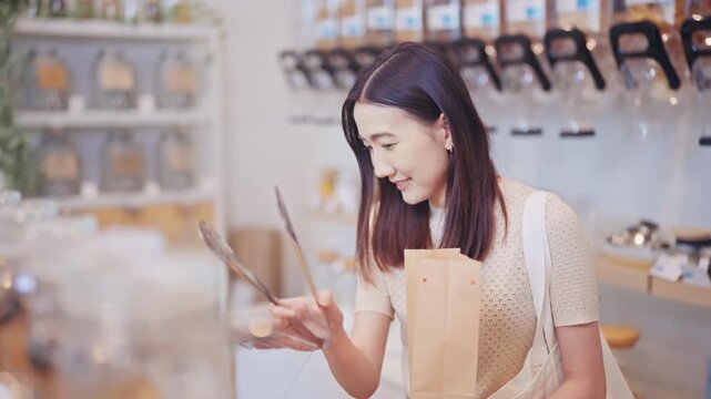 Happy young Asian woman uses tongs to pick products into a brown paper bag, enjoying eco-friendly shopping at a modern refill grocery store with a sustainable lifestyle.