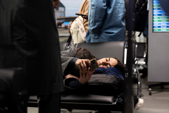 Tired woman laying down on airport bench, texting friends while travelling to foreign country. Fatigued tourist resting on pillow in departure terminal, scrolling on smartphone, waiting to catch plane