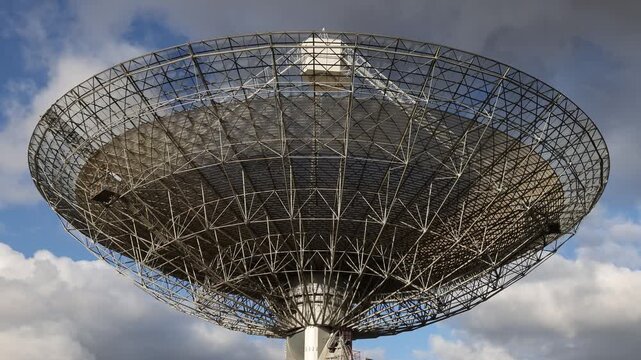 Large satellite dish antenna rotating against cloudy blue sky for space communication.