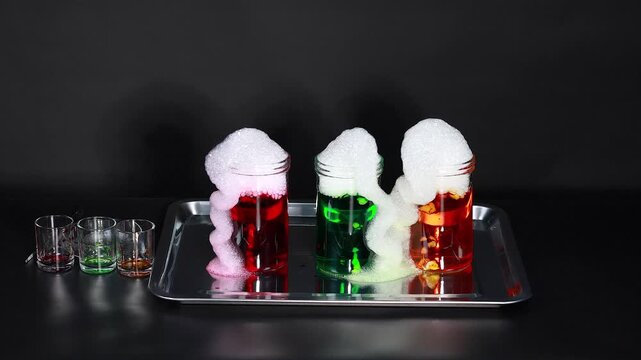 Colored chemical reaction with foam overflowing glass jars on laboratory tray for science experiment.