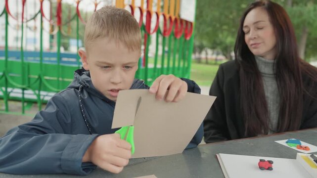 Young boy assembling school craft outdoors. Child carefully cuts cardboard for school project outside. Little boy concentrates on creating craft from cardboard with supervision outside