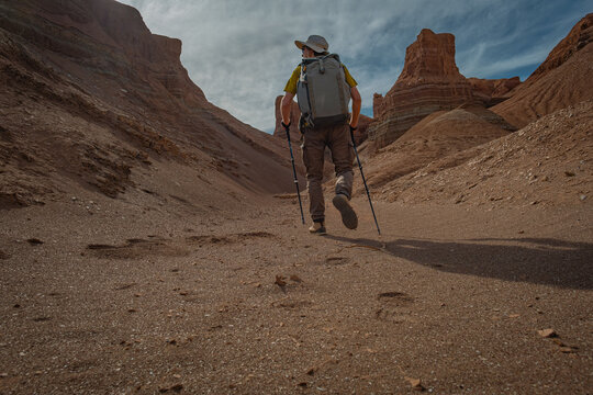Hiker with backpack and trekking poles walking through a desert canyon. Low angle rear view of a traveler exploring red rock formations under a cloudy sky.