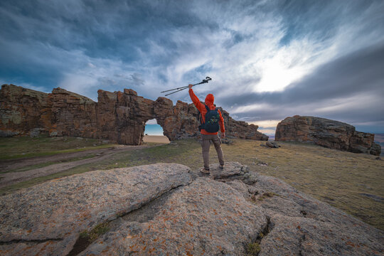 Happy hiker raising trekking poles in a victory gesture standing on a rock near a natural stone arch. Adventure, success and achievement concept in the wilderness.