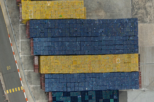 Aerial view of rows of blue and yellow plastic crates stacked in an industrial logistics yard next to a paved road with markings.