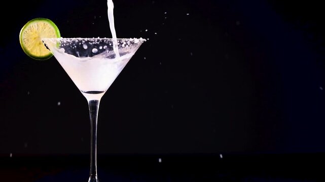 Slow motion pouring white margarita cocktail into salt rimmed glass with lime on black background.