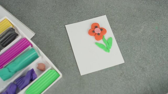 Colorful clay flower on white card with modeling kit nearby, neat craft tray and vibrant petals on classroom table, finished art project ready for school display or take home, tidy supplies suggesting