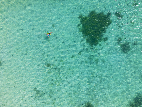 Aerial view of a person floating in the crystal clear turquoise sea near a dark coral reef patch in Guanaja, Bay Islands Department, Honduras.