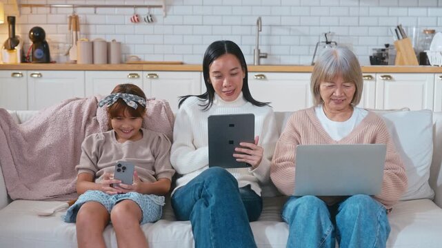 Asian three generations family sits together on a sofa. The young girl is playing on a mobile phone, the mother is working on a tablet, and the grandmother is using a laptop in a cozy, modern home