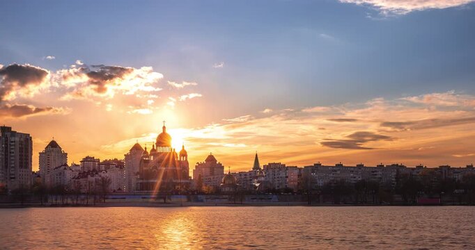 Sunset over Kyiv skyline on the Dnipro River embankment, golden light behind cathedral domes, reflections on water and dramatic clouds creating a calm urban evening atmosphere. Time lapse